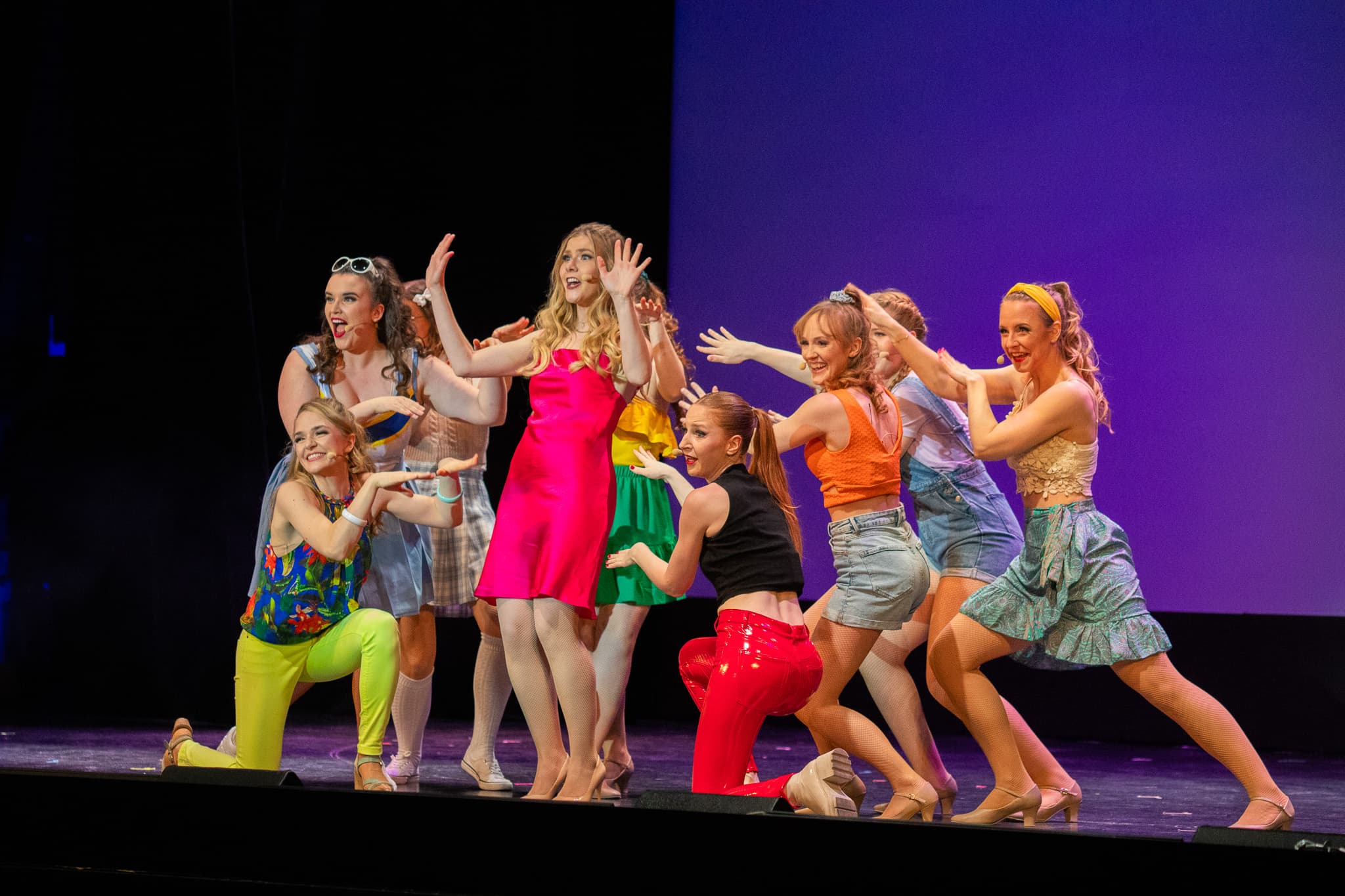 Frau in pinkem Kleid steht im Mittelpunkt, Ensemble zeigt gestische Bewegungen und richtet Aufmerksamkeit auf sie.