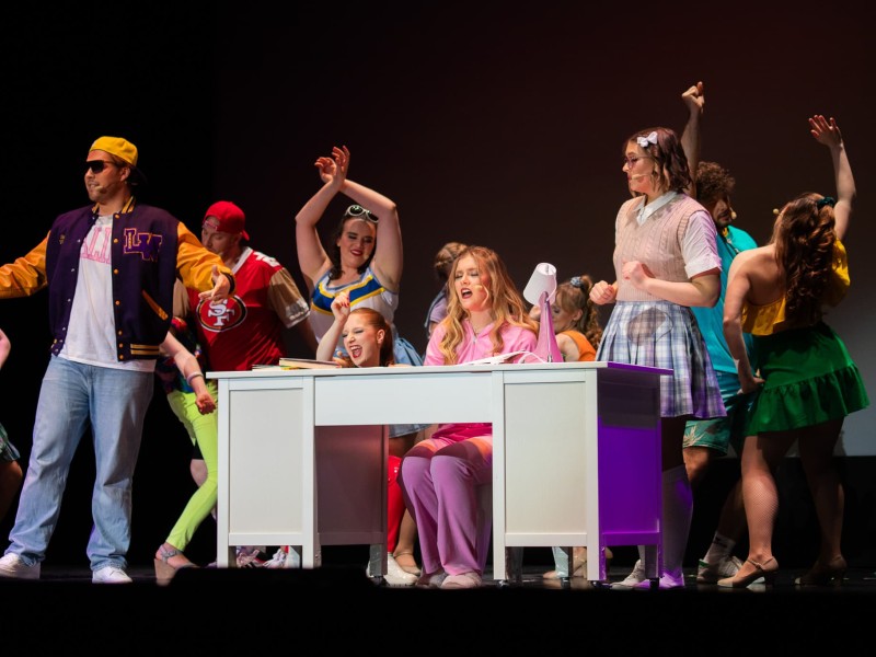 Ensemble tanzt um Schreibtisch, eine Frau sitzt daran, andere bewegen sich dynamisch in bunten Kostümen.