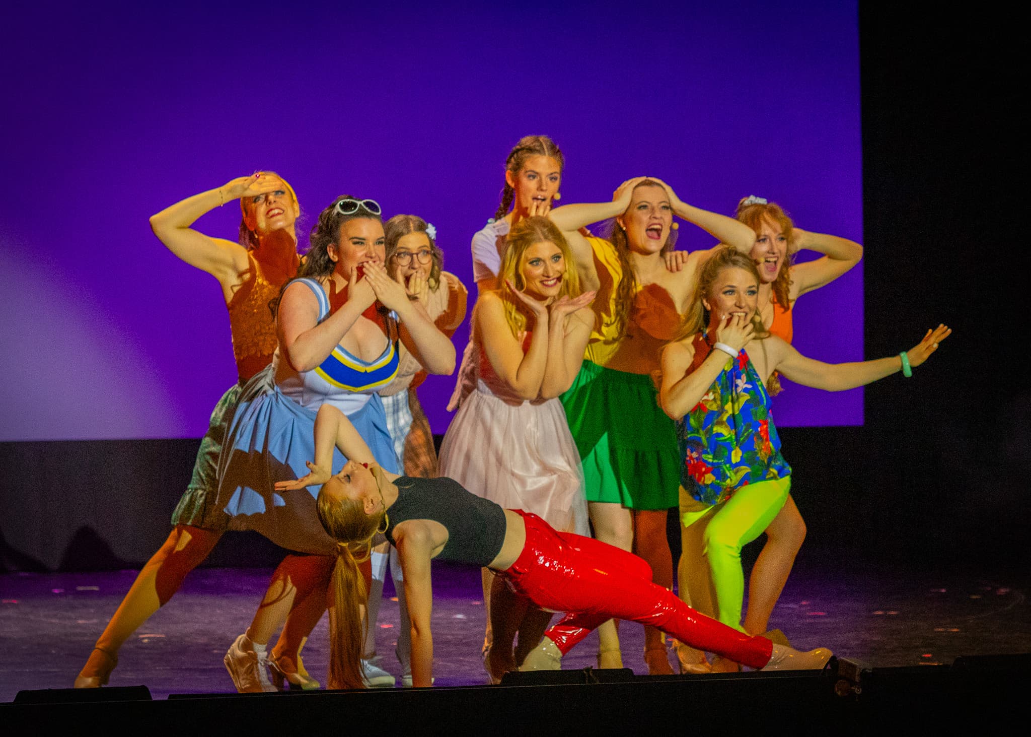 Tanzensemble auf Bühne in bunten Kostümen, mehrere Frauen posieren überrascht und gestikulierend