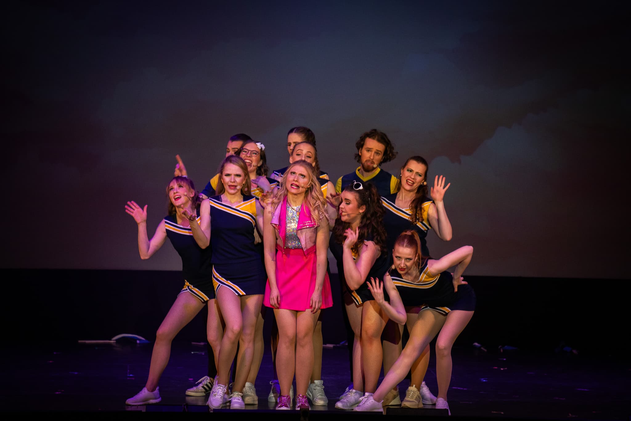 Gruppe in Cheerleader-Outfits steht eng zusammen, eine Frau in pink steht im Zentrum mit unsicherem Ausdruck.