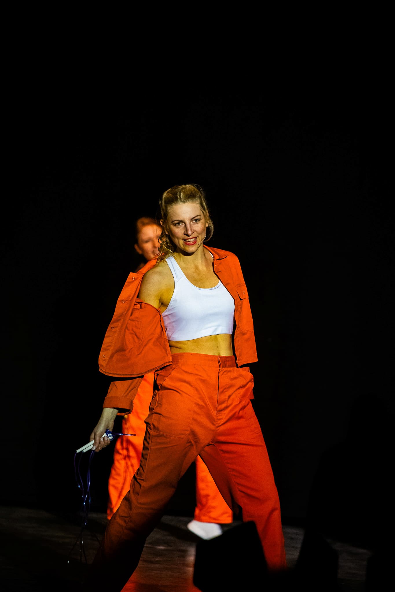 Darstellerin in orangefarbener Sportkleidung hält Springseil und bewegt sich auf dunkler Bühne