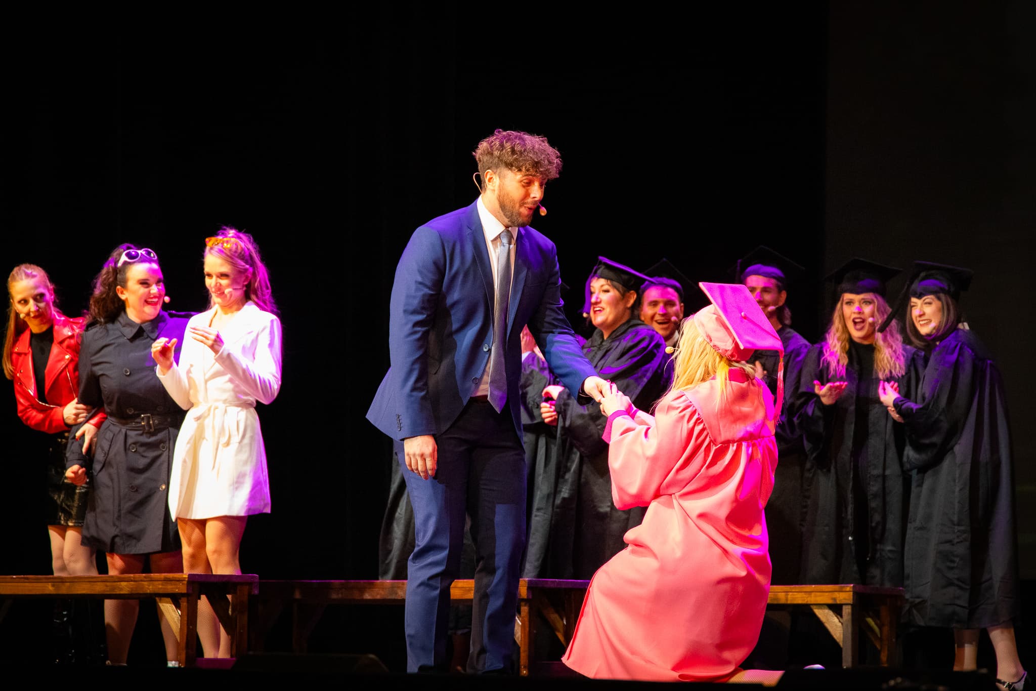 Mann im Anzug hilft Frau in pinker Abschlussrobe auf, Gruppe im Hintergrund applaudiert.