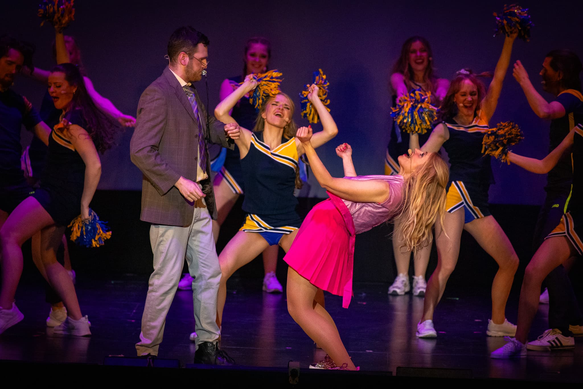 Cheerleader tanzen im Hintergrund, Frau in pink lehnt sich zurück. Ein Mann steht schockiert in der Mitte.
