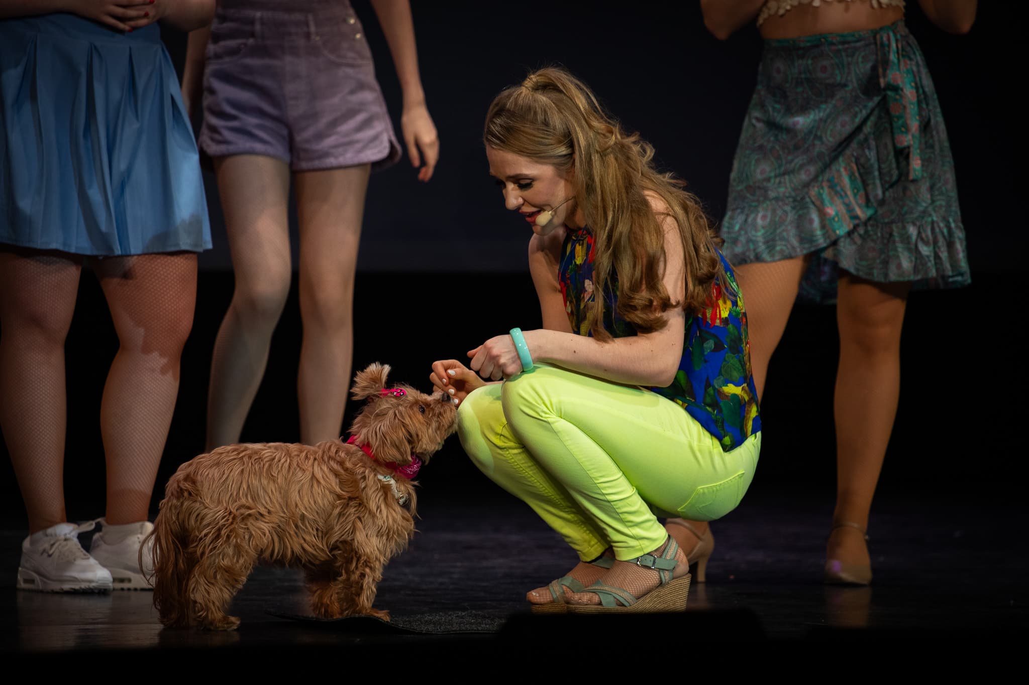 Frau kniet auf der Bühne und füttert einen kleinen Hund mit Leckerli, während im Hintergrund weitere Darstellerinnen stehen