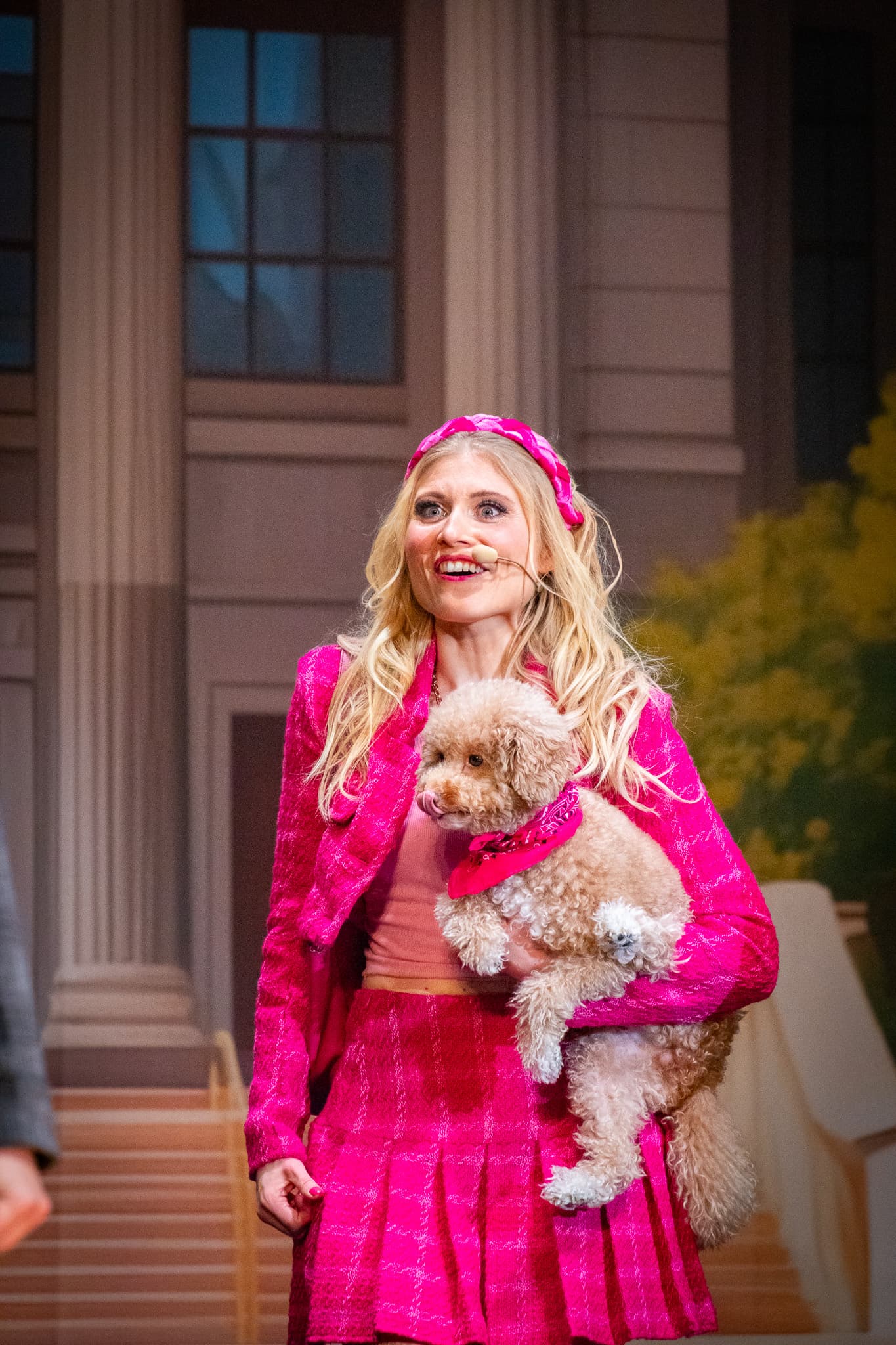 Frau in pinkem Kostüm hält kleinen Hund im Arm, steht auf Bühne vor Gebäudekulisse