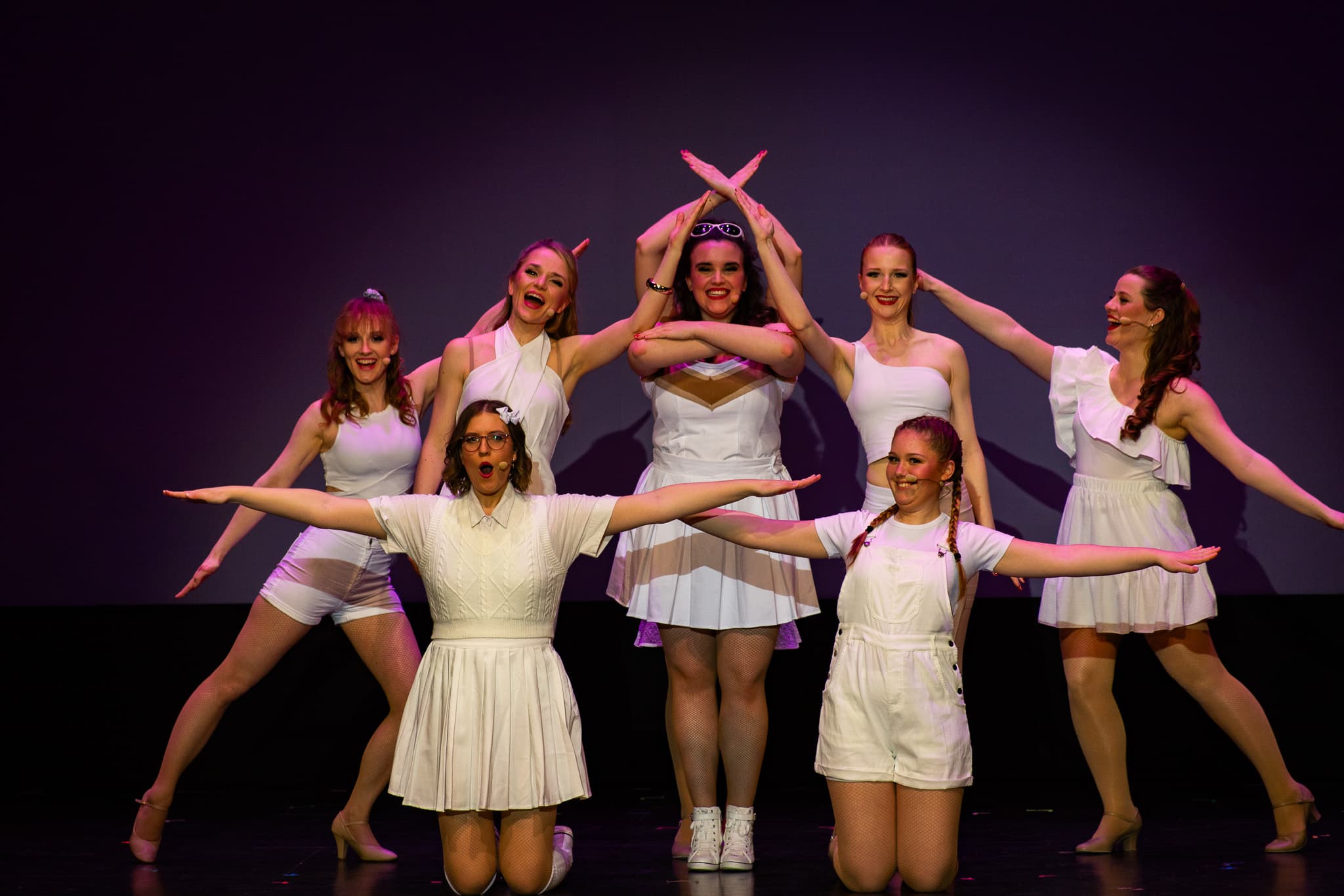 Tanzgruppe in weißen Outfits posiert symmetrisch auf Bühne, Arme ausgestreckt, lächelnde Gesichter.