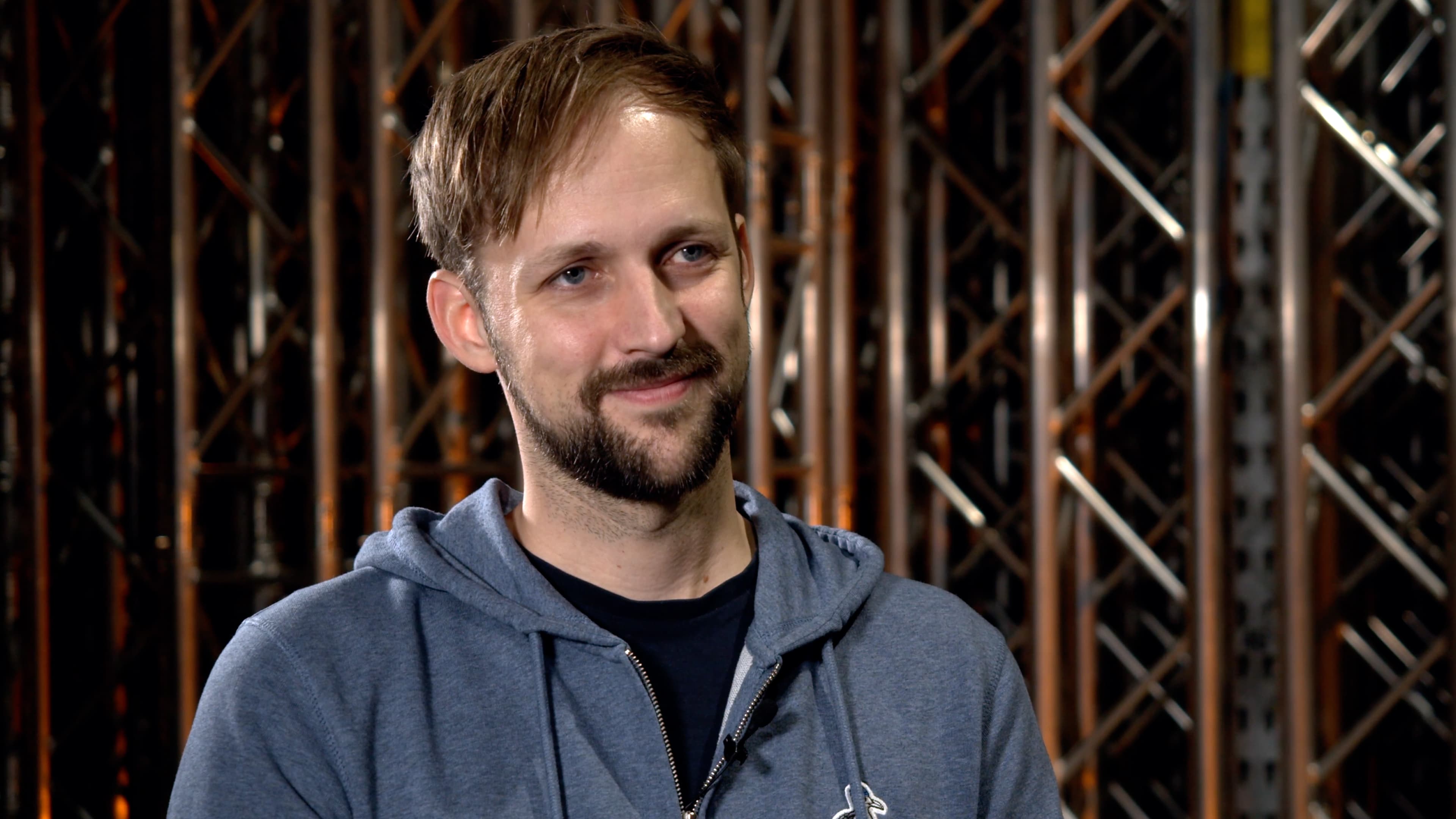 Mann mit Bart in grauem Hoodie sitzt vor Bühnenkonstruktion und blickt zur Seite