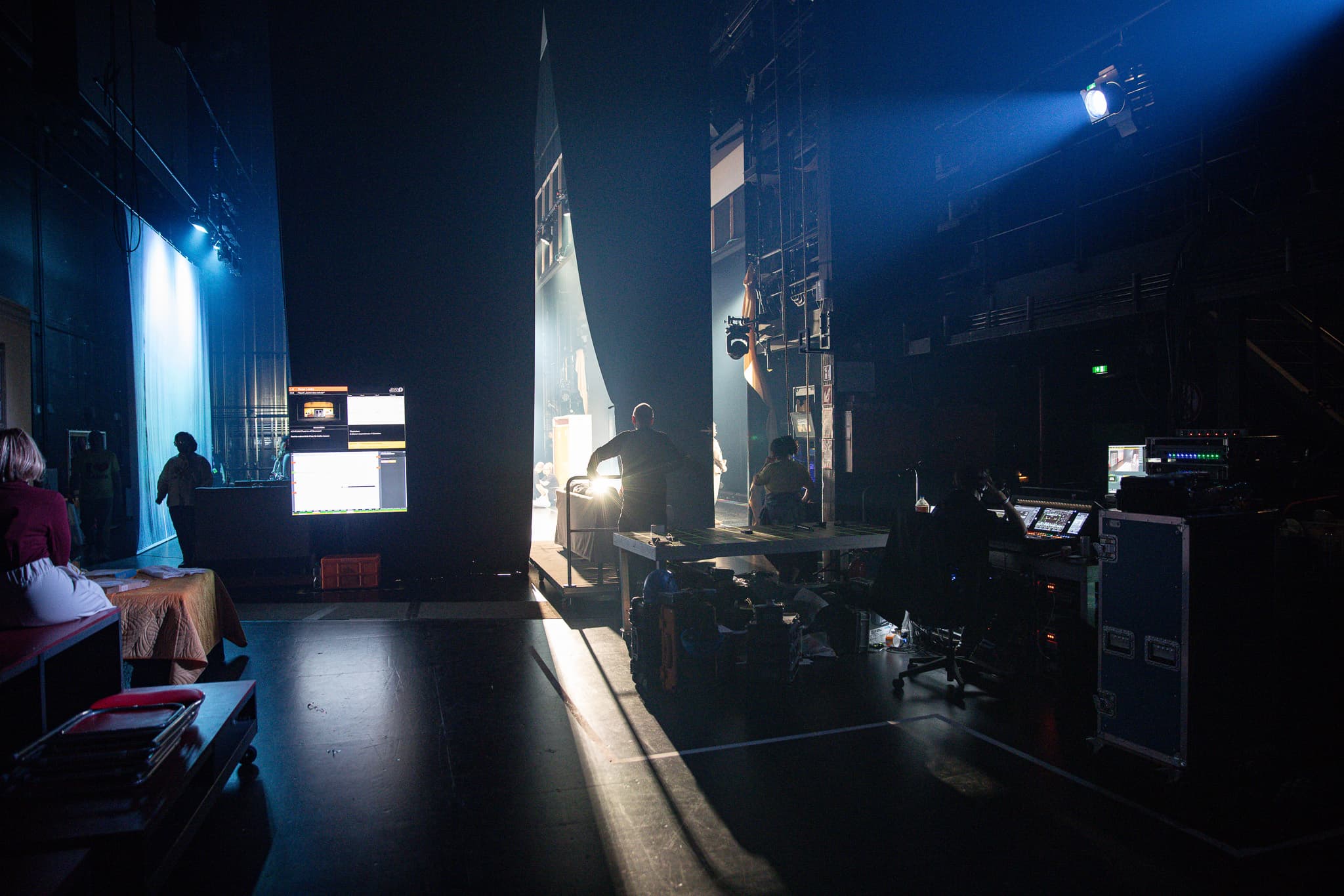 Blick hinter die Kulissen einer Theaterbühne mit technischem Equipment, Monitoren und Personal im Halbdunkel.