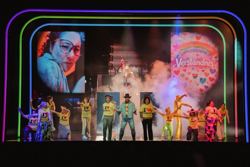 Eine Theaterbühne mit vielen Darstellern, gerahmt von Neonbögen und zwei Bildschirmen, wobei eine Person im Zentrum im Nebel an Seilen nach oben gezogen wird.