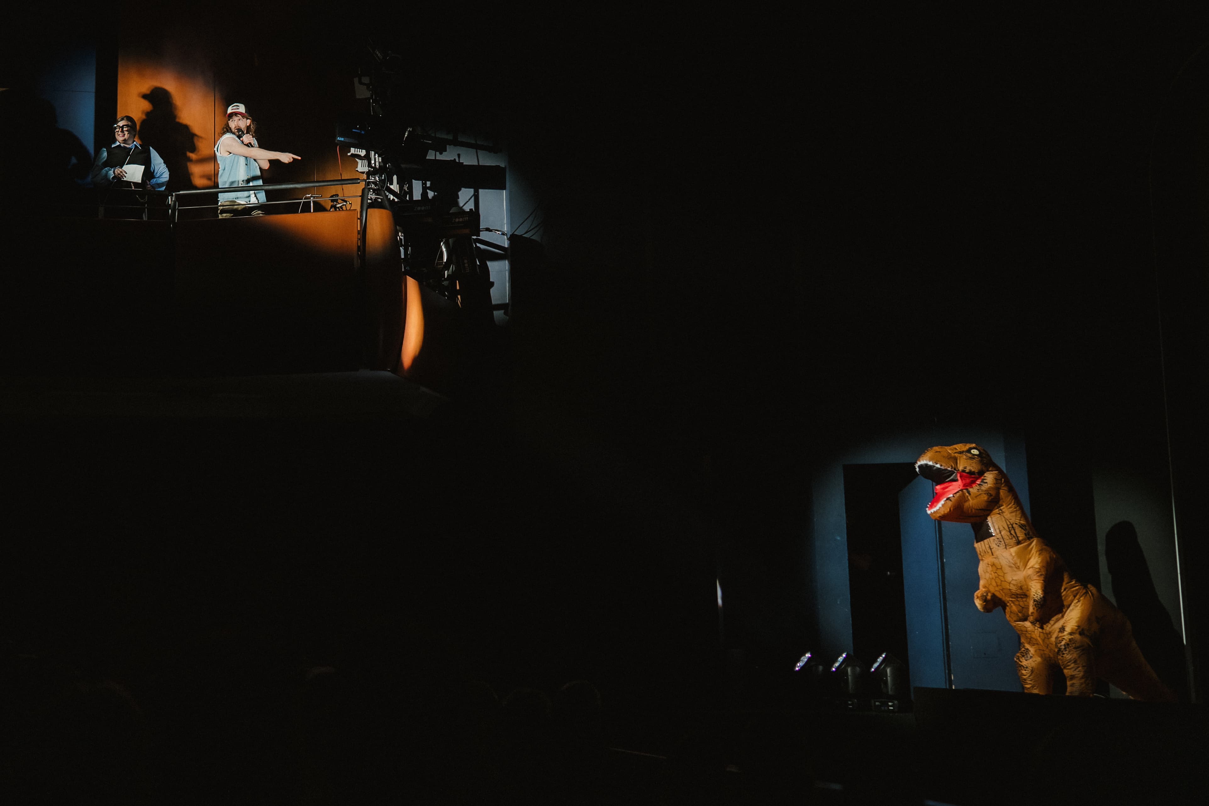 Eine Person in einem aufblasbaren Dinosaurier-Kostüm steht auf einer Bühne im Scheinwerferlicht, während zwei Personen von einem Balkon aus auf sie zeigen.