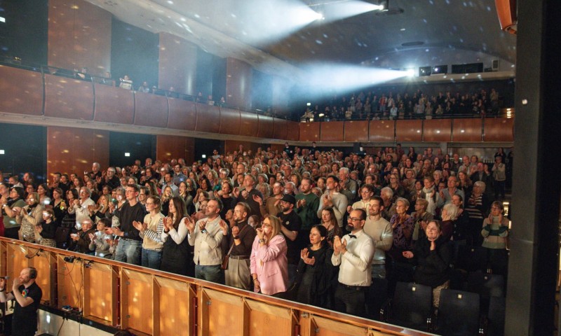 Ein dicht besetzter Theatersaal mit Publikum, das stehend applaudiert, während Scheinwerferkegel durch den dunklen Raum leuchten.