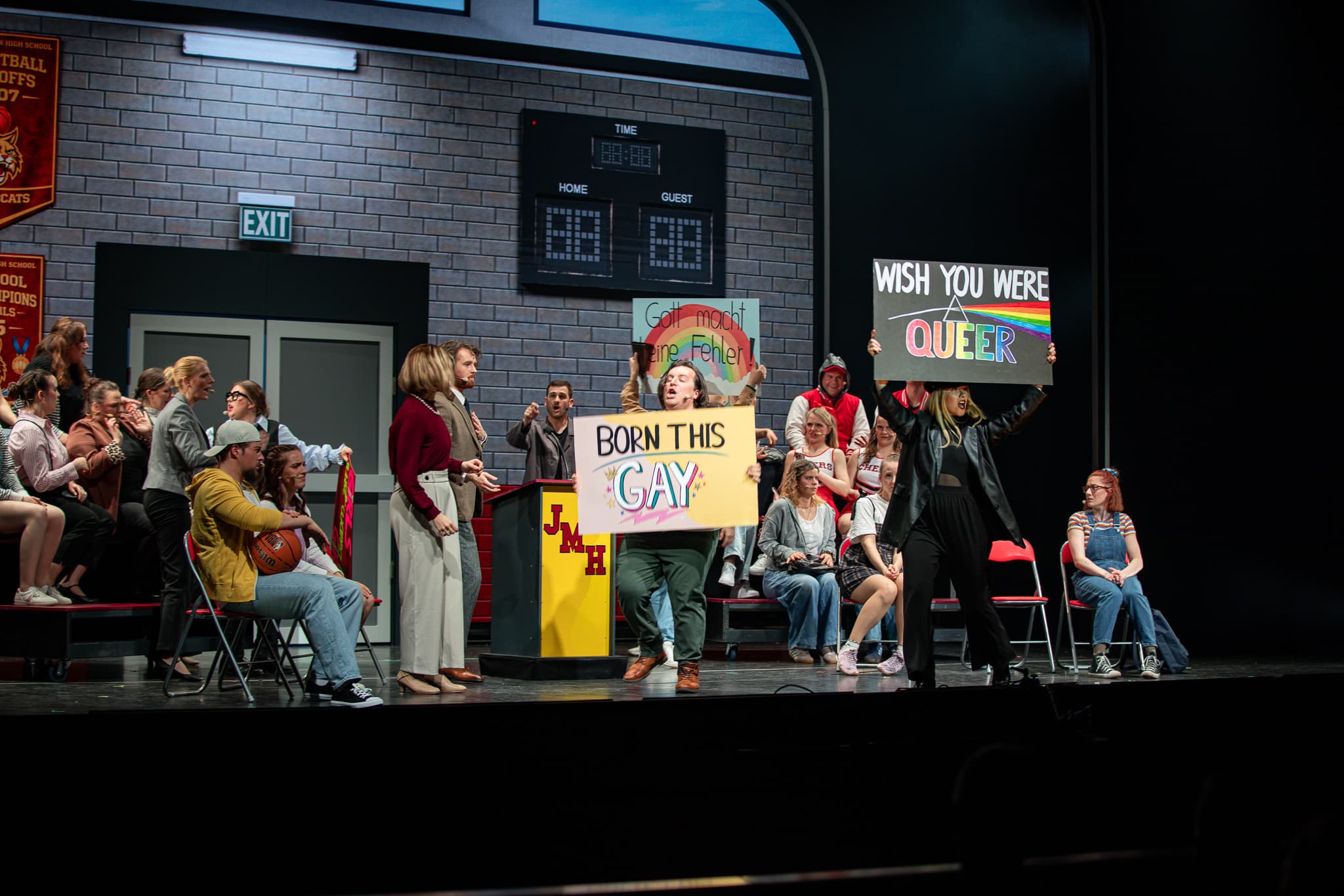 Theaterszene mit Demonstration: Personen halten Schilder mit Botschaften wie "Born this Gay" und "Wish you were queer"