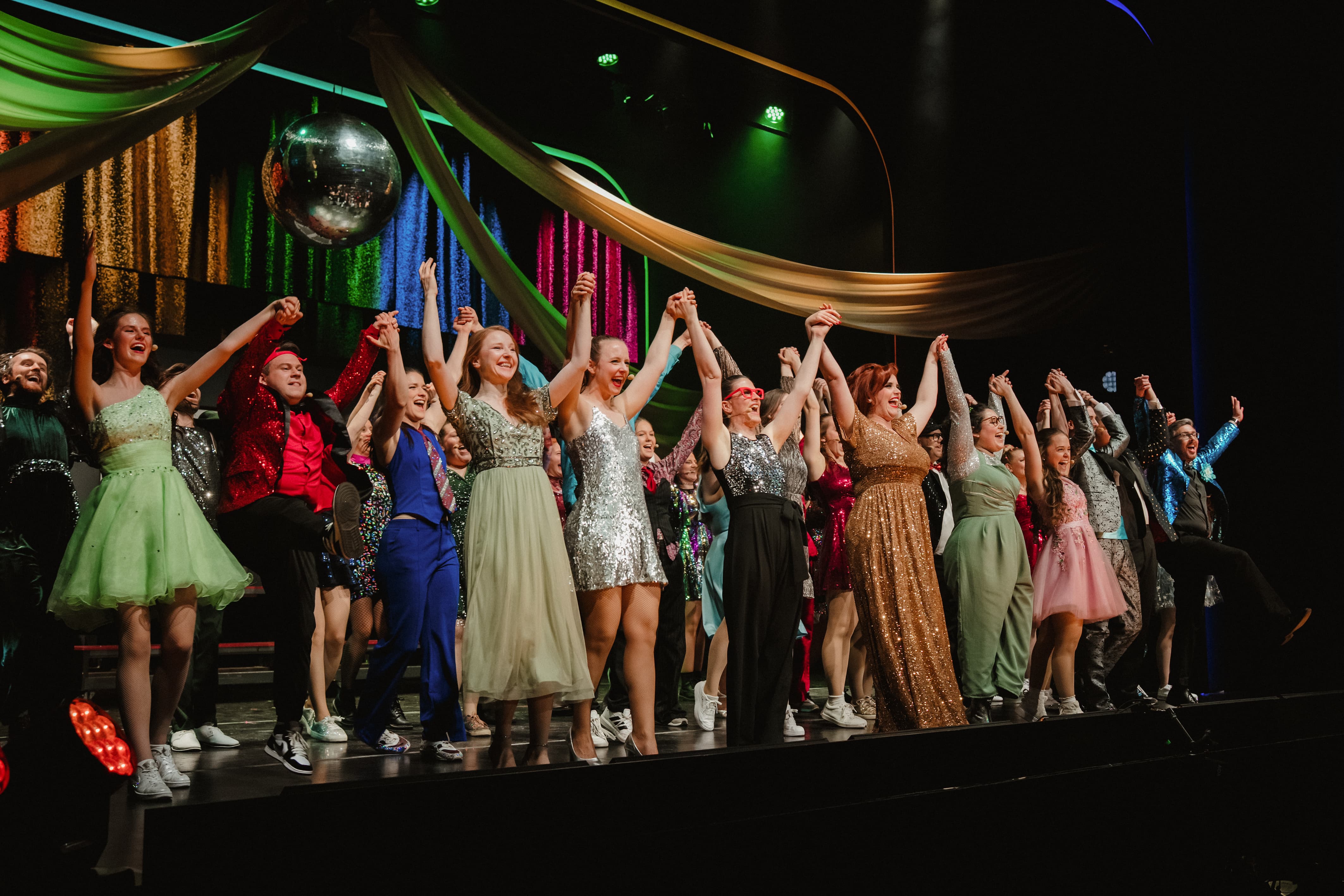 Eine große Gruppe von Darstellern in festlicher, glitzernder Kleidung steht jubelnd mit erhobenen, gefassten Händen auf einer Theaterbühne unter einer Diskokugel.