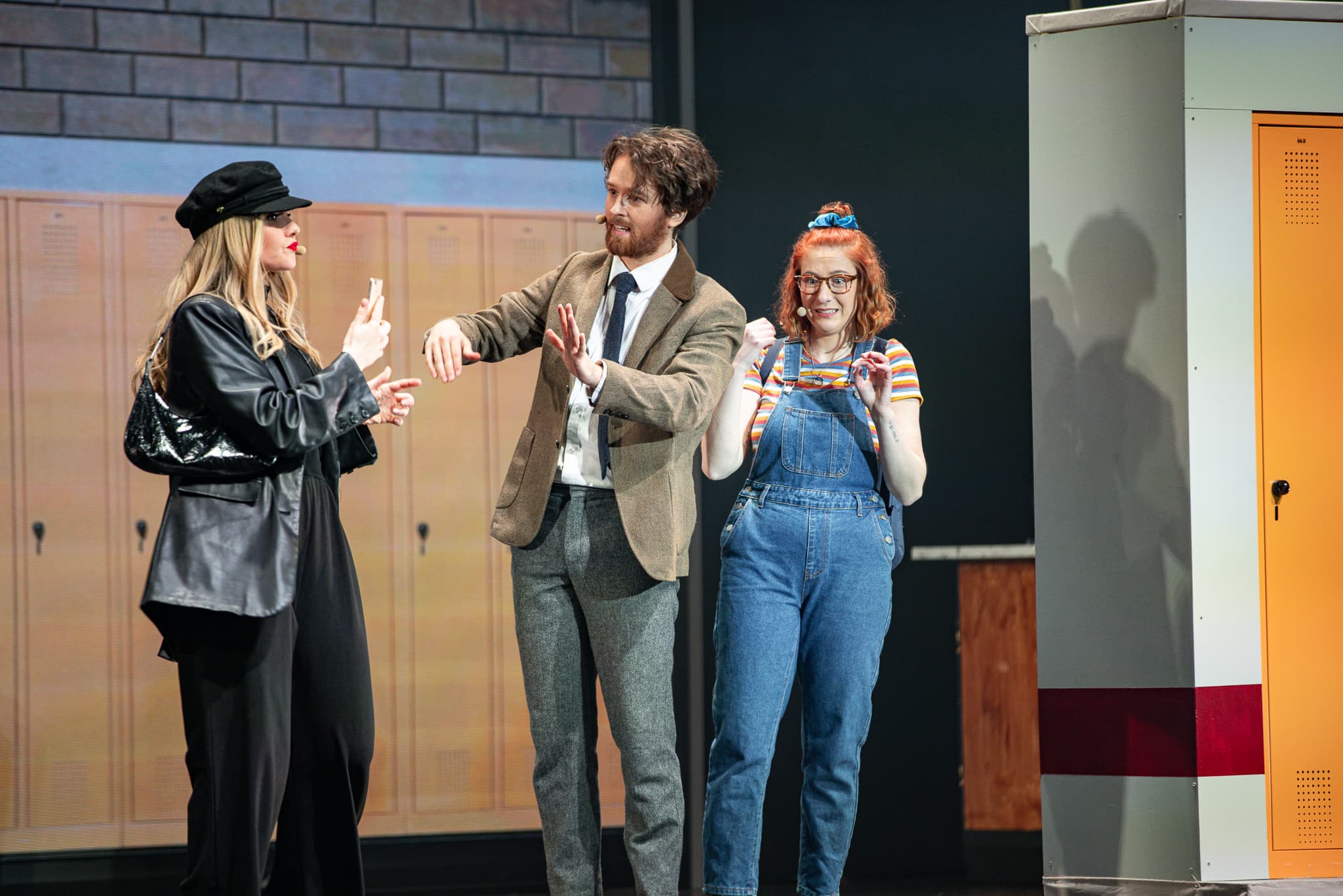 Drei Personen in auffälligen Kostümen agieren vor einer Kulisse aus Schulspinden auf einer Theaterbühne.