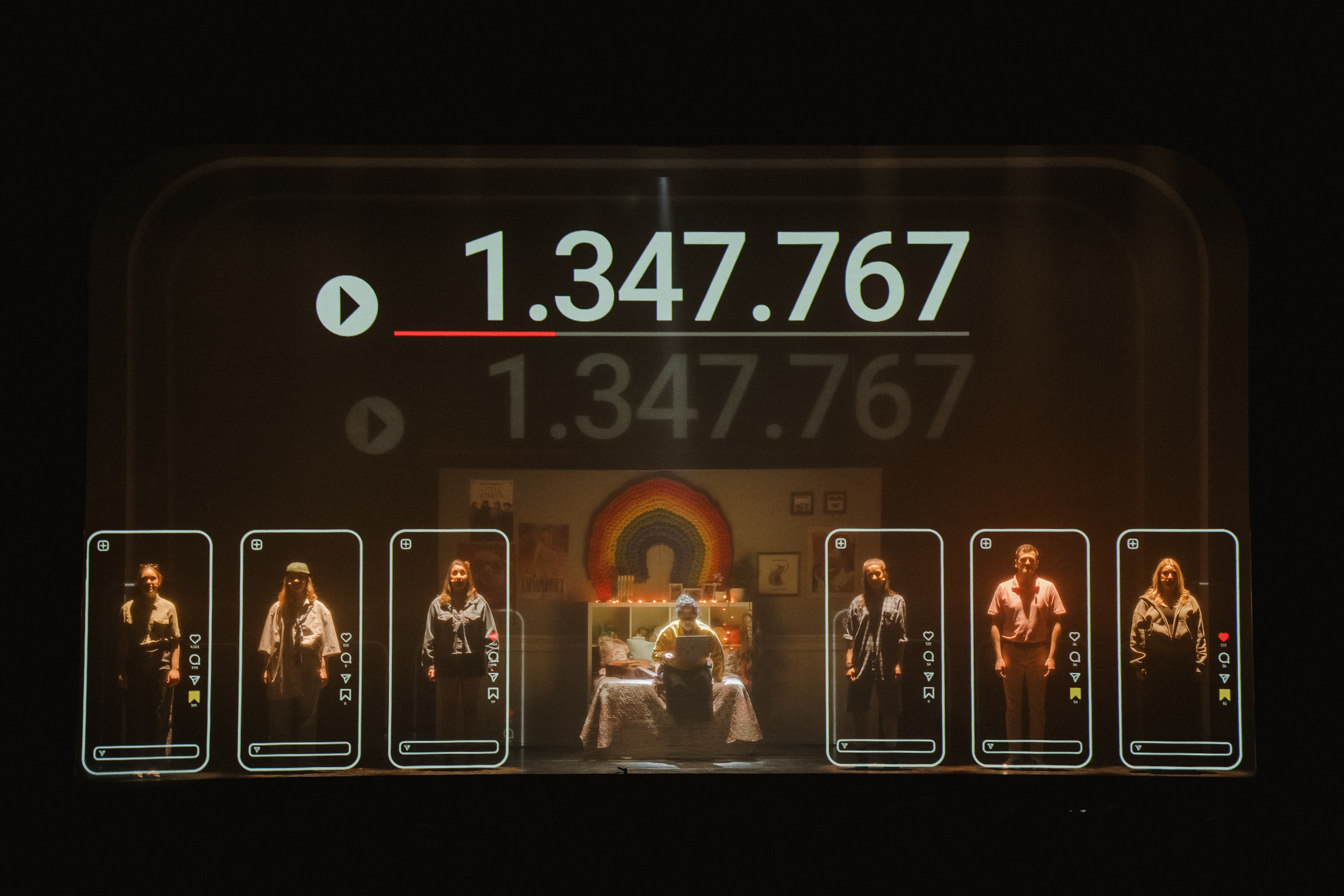 Eine Theaterbühne mit einer großen Projektion der Zahl 1.347.767 und sechs Schauspielern, die einzeln in leuchtenden Smartphone-Rahmen stehen.
