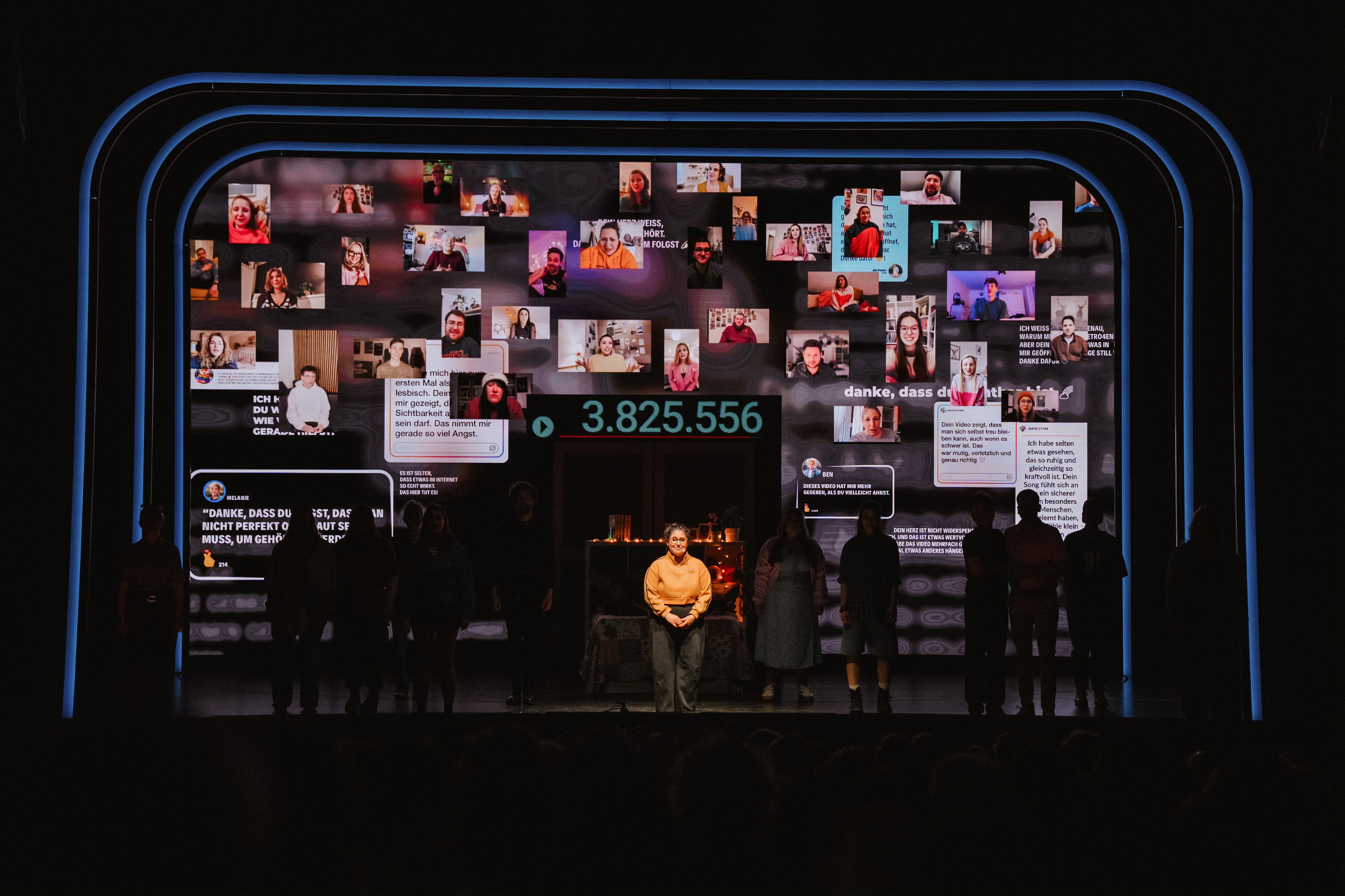 Eine Theaterbühne mit einer großen Videoprojektion aus zahlreichen Porträt-Kacheln, Textnachrichten und der Zahl 3.825.556, vor der eine einzelne Person im Scheinwerferlicht steht.