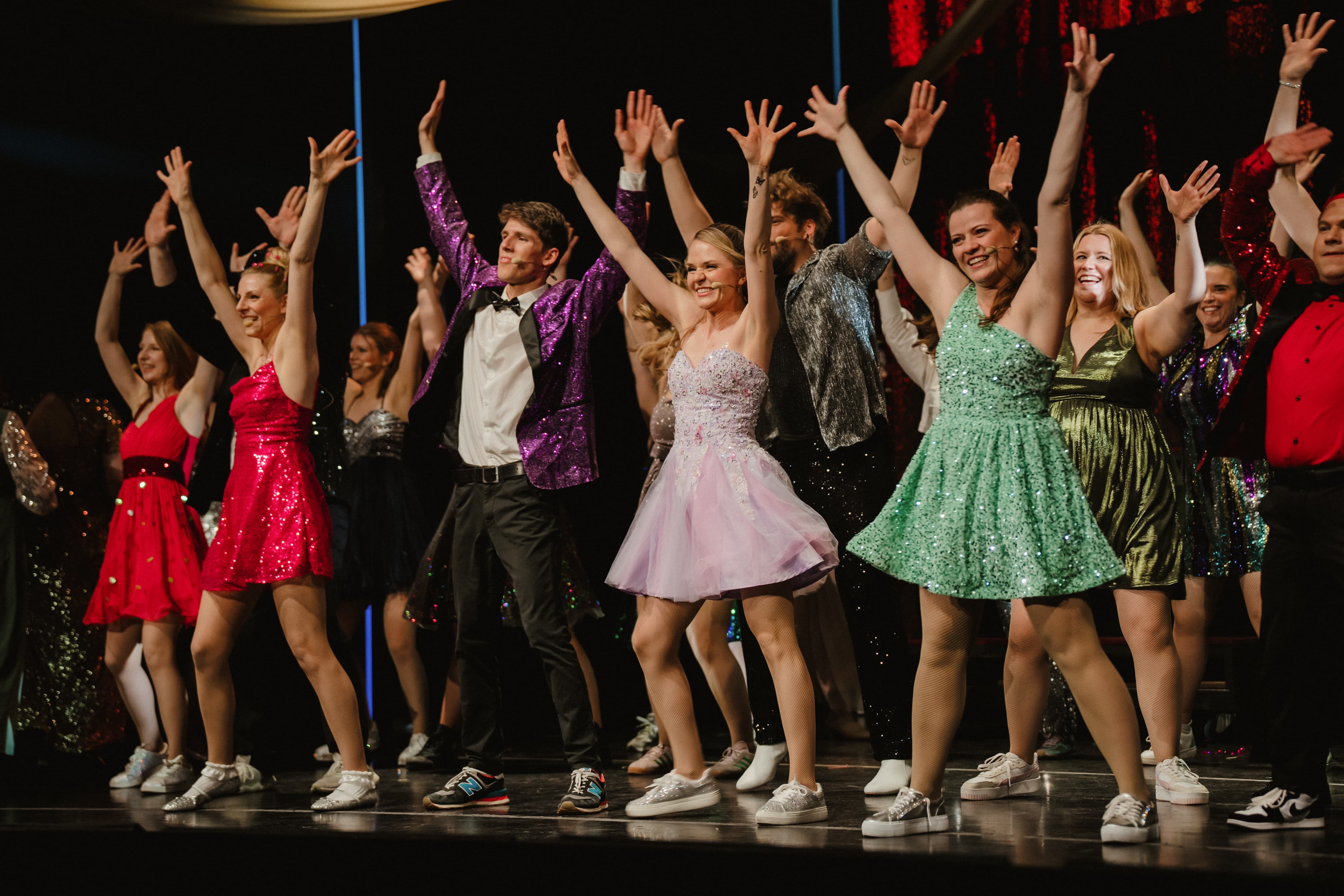 Ein Musical-Ensemble in glitzernder Abendgarderobe jubelt mit nach oben gestreckten Armen auf einer Theaterbühne.
