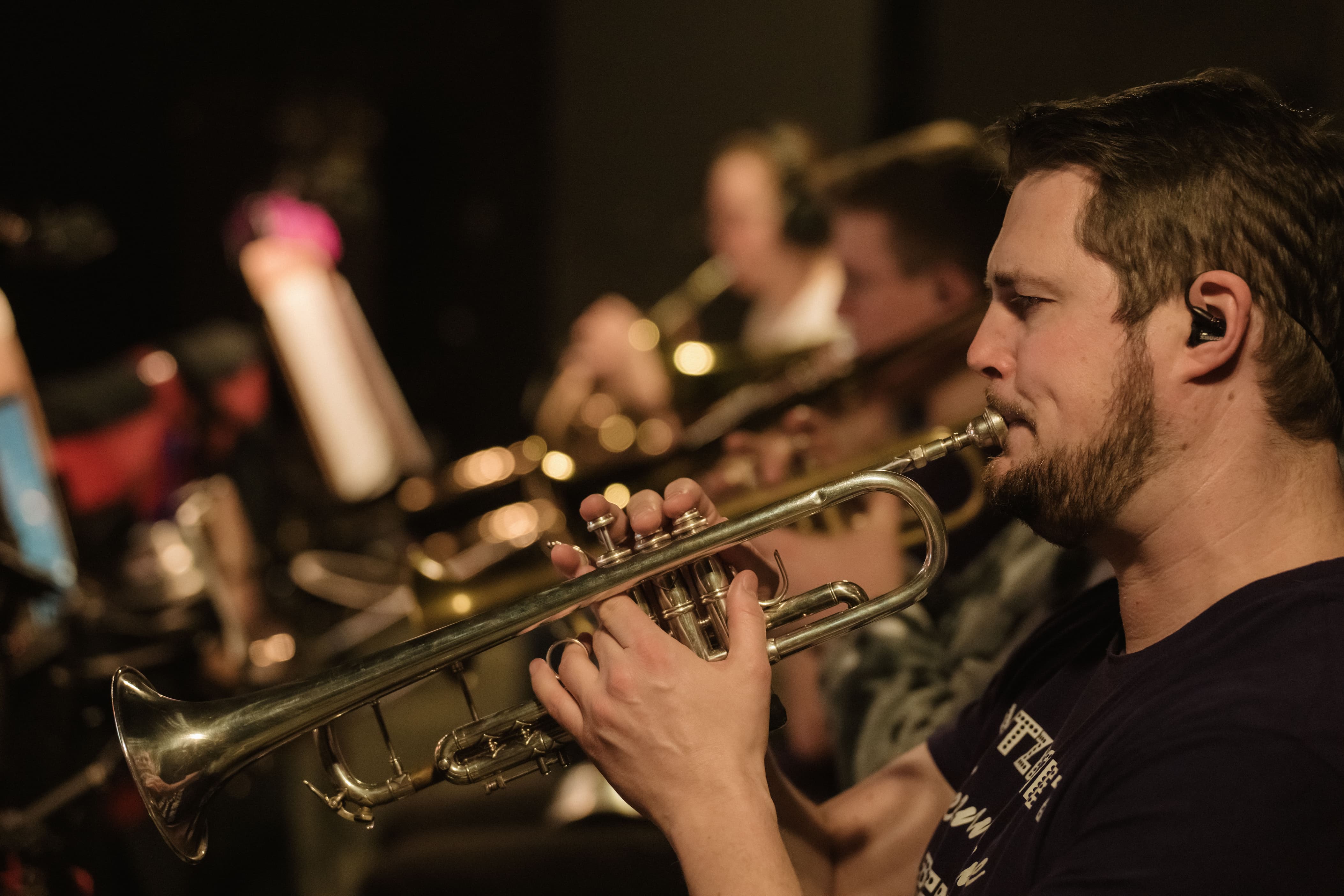 Eine Gruppe von Musiker*innen spielt in einer schwach beleuchteten Umgebung Blechblasinstrumente, wobei ein Mann im Vordergrund die Trompete spielt.