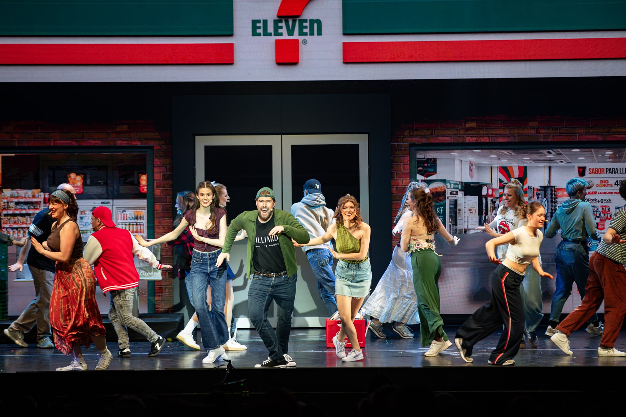 Ensemble tanzt vor einer Kulisse mit einem Laden im Stil eines 7-Eleven, mehrere Personen bewegen sich in unterschiedliche Richtungen.