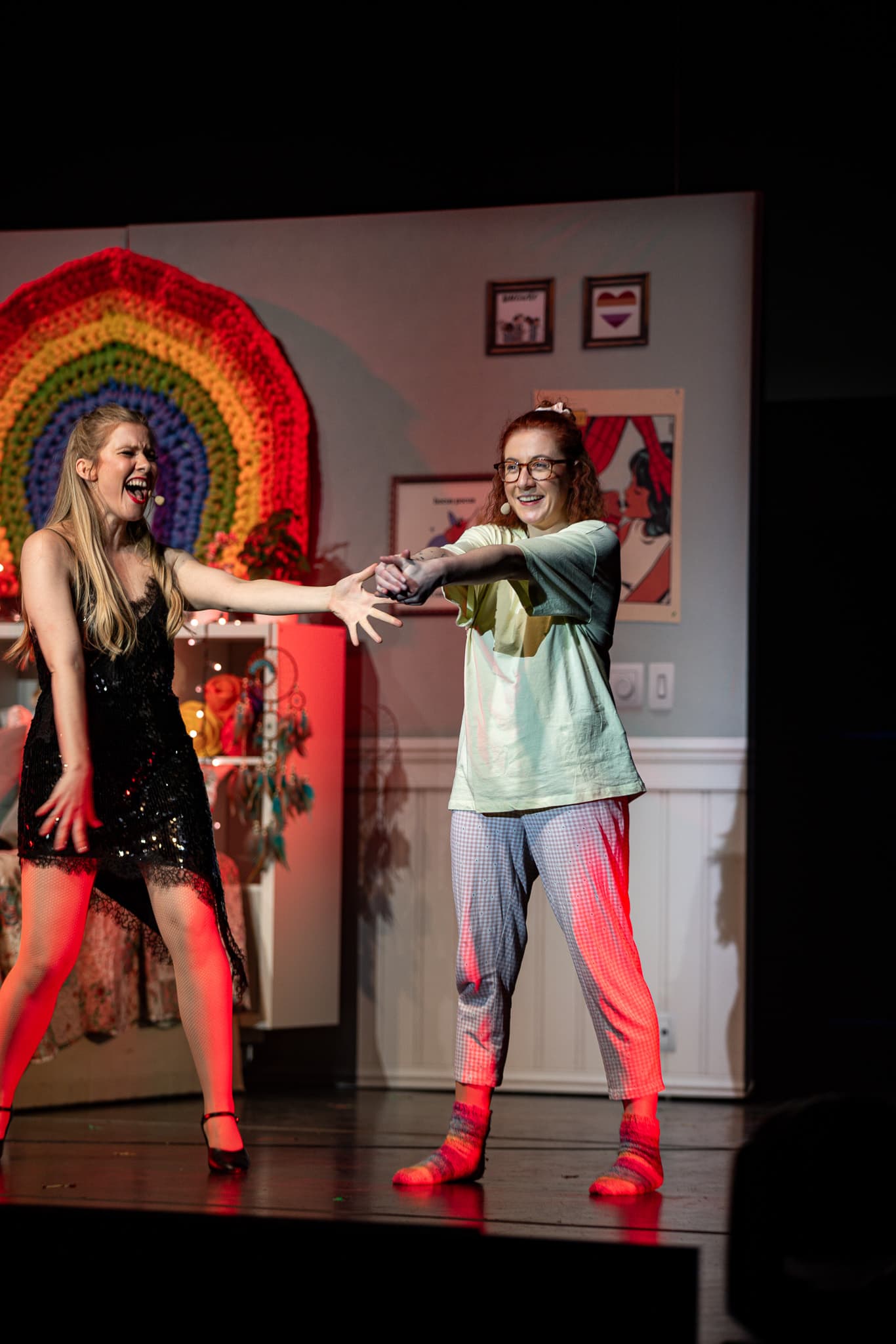 Zwei Frauen halten sich an den Händen und spielen eine emotionale Szene vor einer Regenbogen-Dekoration.