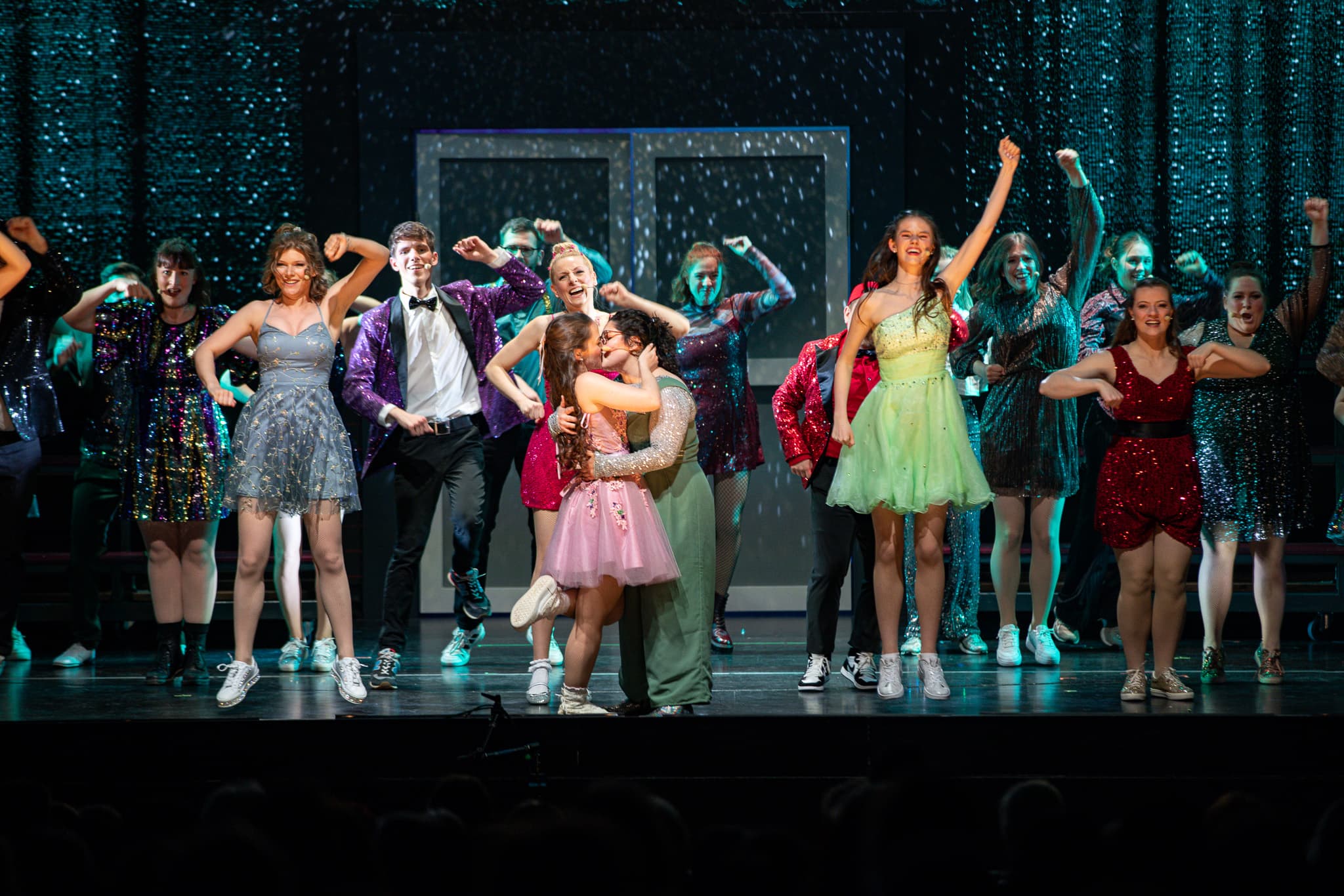 Bunte Musicalszene auf einer Bühne: Eine Gruppe von Jugendlichen in glitzernden Kostümen tanzt, während zwei Mädchen im Vordergrund sich küssen und vom Ensemble jubelnd umgeben sind.