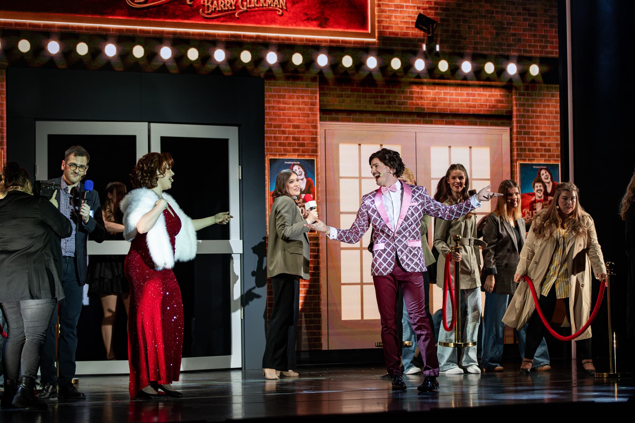 Bühnenszene mit Moderator und Darstellenden: Eine Frau im roten Kleid und ein Mann im auffälligen Anzug stehen im Mittelpunkt.