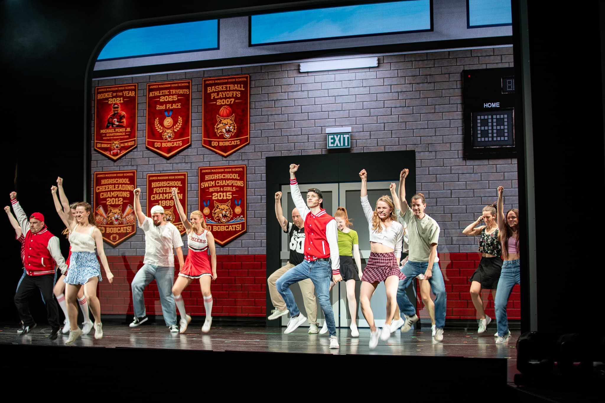 Tanzszene in einer Schulkulisse: Gruppe von Jugendlichen springt synchron mit erhobenen Armen auf der Bühne.