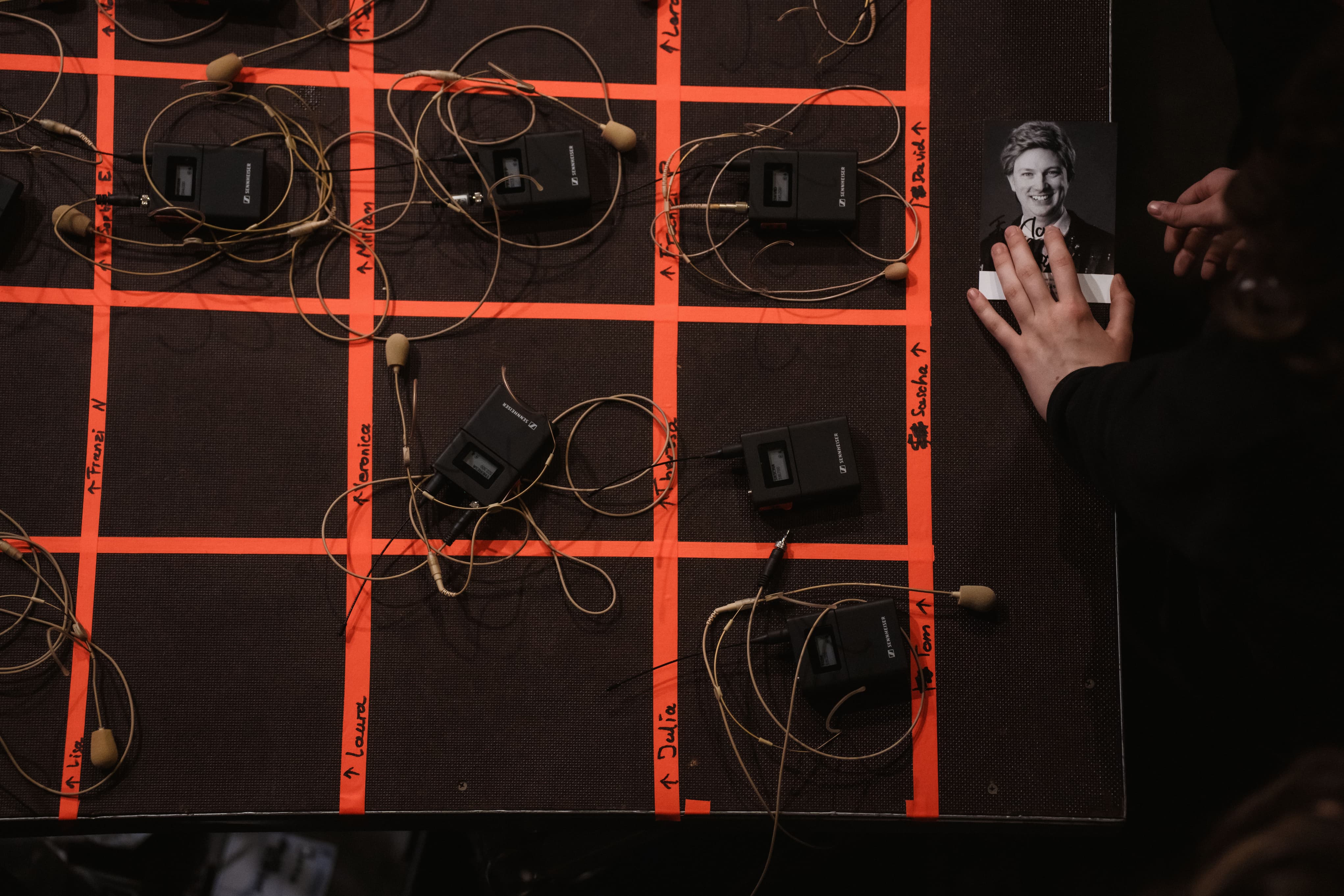 Mehrere drahtlose Mikrofone und sind in einem mit orangefarbenem Klebeband markierten Raster angeordnet, während eine Hand ein Schwarz-Weiß-Foto einer Person auf der rechten Seite des Tisches hält.