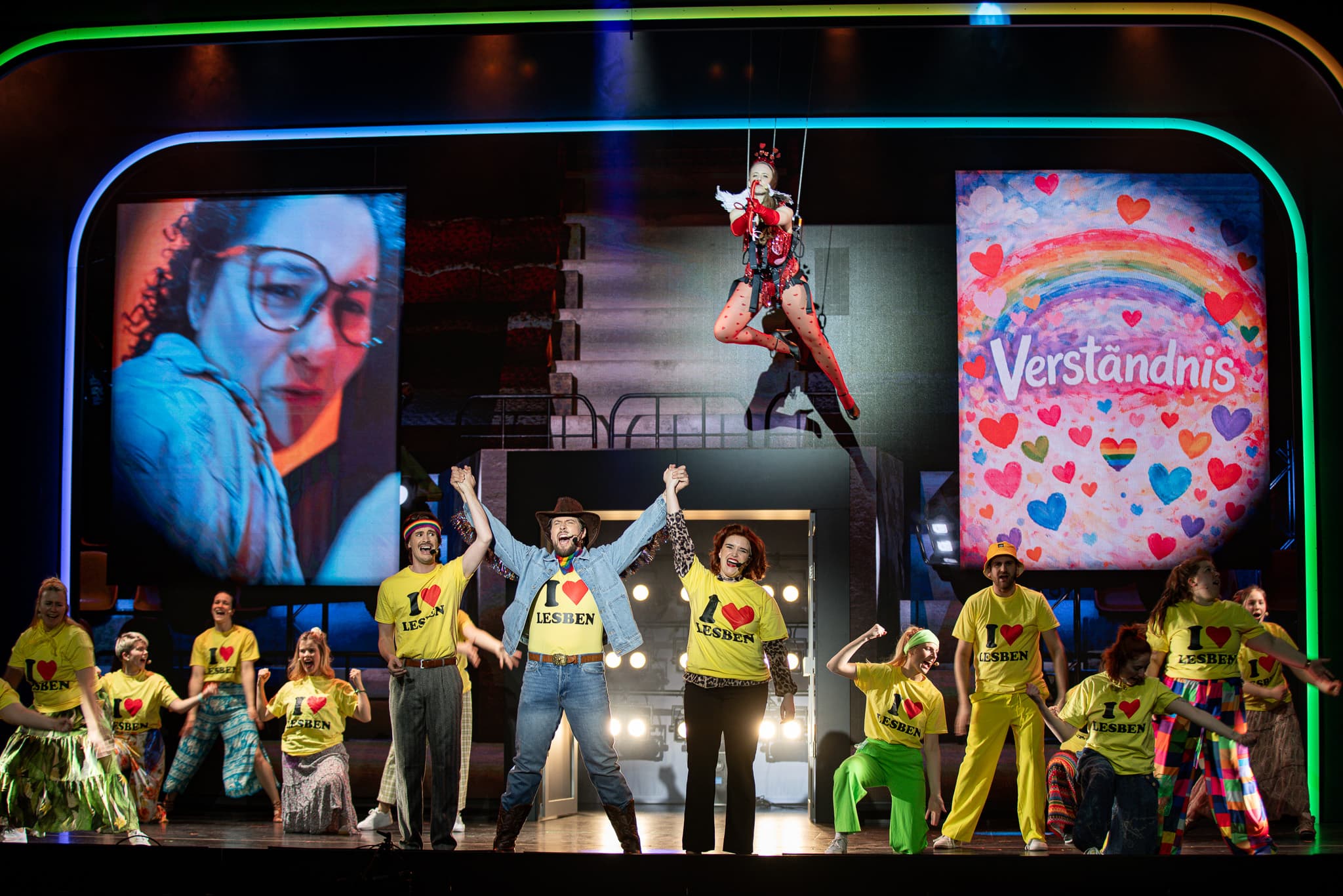 Großes Ensemble in gelben T-Shirts mit Aufschrift "I love Lesben" tanzt vor bunten Projektionen mit Regenbogenmotiv.