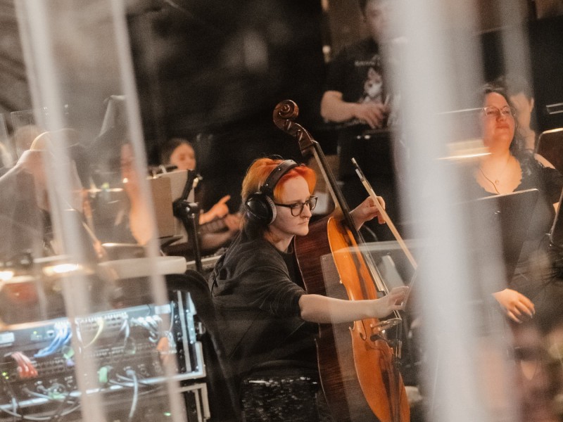 Eine junge Person mit Brille, Kopfhörern und orangefarbenem Haar sitzt konzentriert an einem Cello inmitten von technischem Equipment.