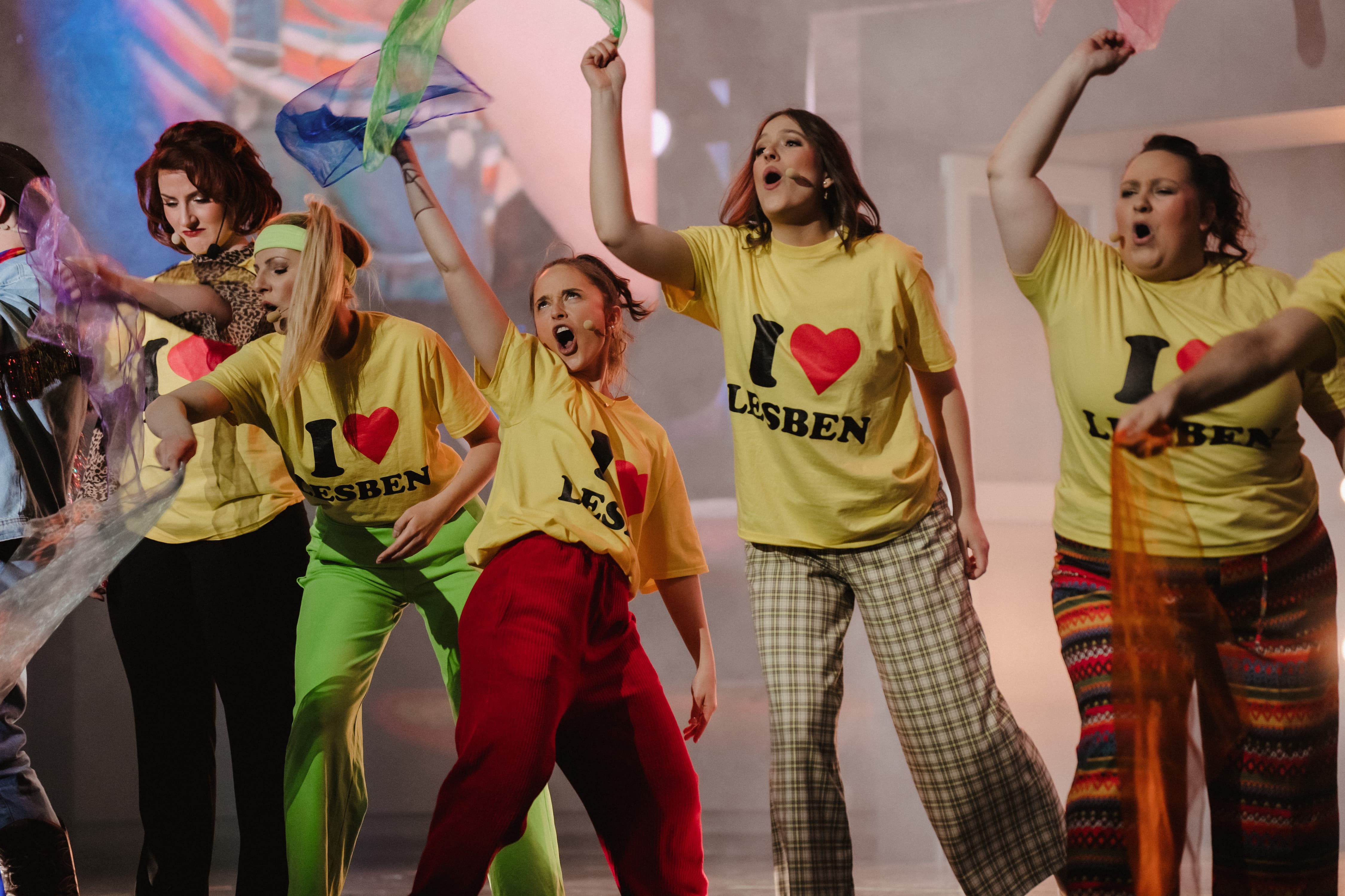 Eine Gruppe von Schauspielerinnen singt und tanzt energisch in bunten Hosen und T-Shirts mit der Aufschrift "I love Lesben", während sie bunte Tücher in die Luft schwingen.