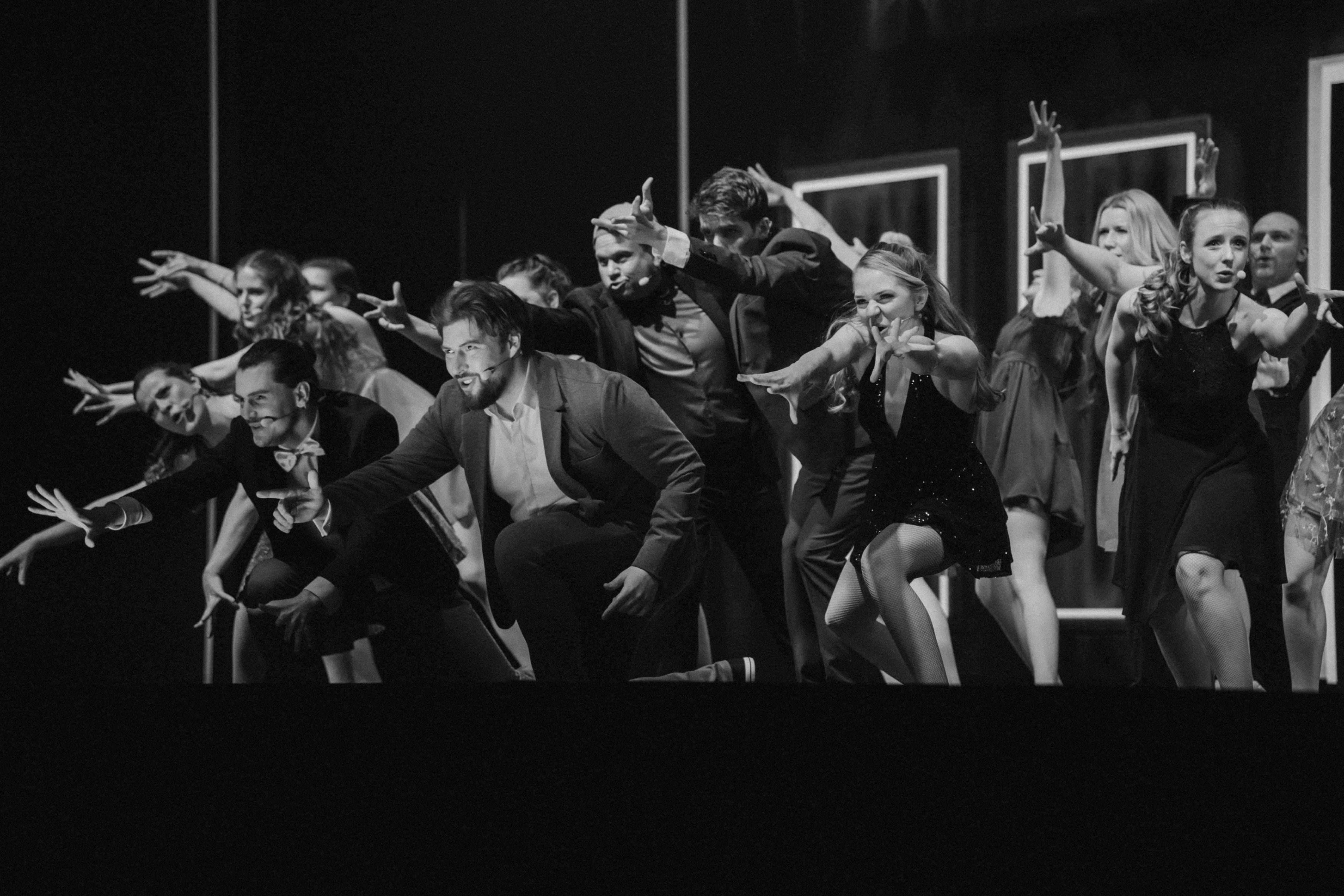 Darsteller einer Theaterproduktion in Anzügen und Kleidern posieren in einer gestaffelten Formation mit weit ausgestreckten Armen und fokussierten Blickrichtungen.
