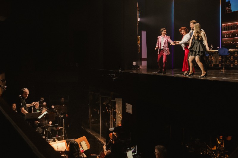 Blick aus dem Zuschauerraum auf eine Theaterbühne mit vier Darstellern in festlicher Kleidung und das tiefer gelegene Orchester im Orchestergraben.