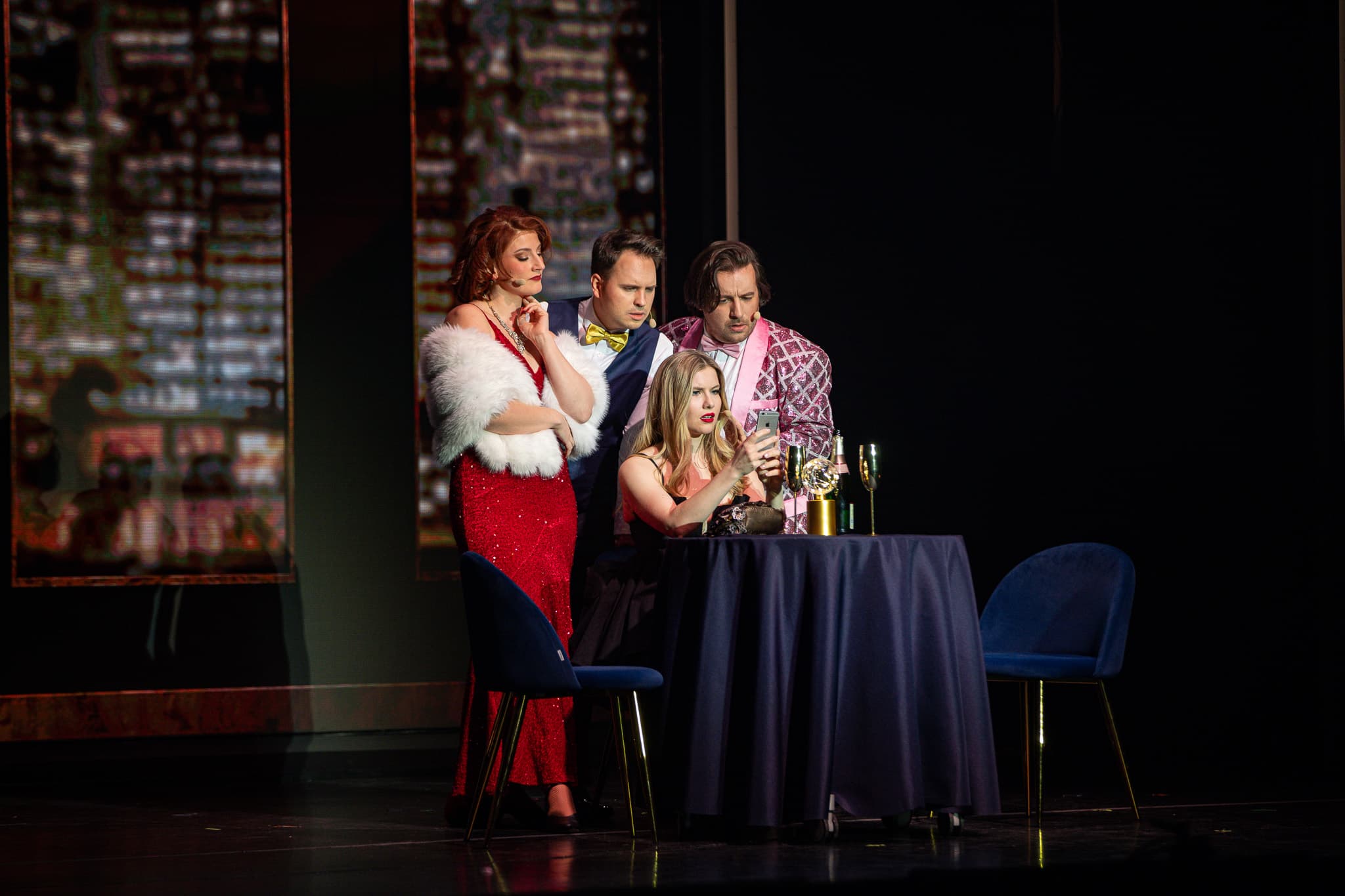 Vier elegant gekleidete Personen auf einer Theaterbühne blicken gespannt und besorgt auf ein Smartphone.