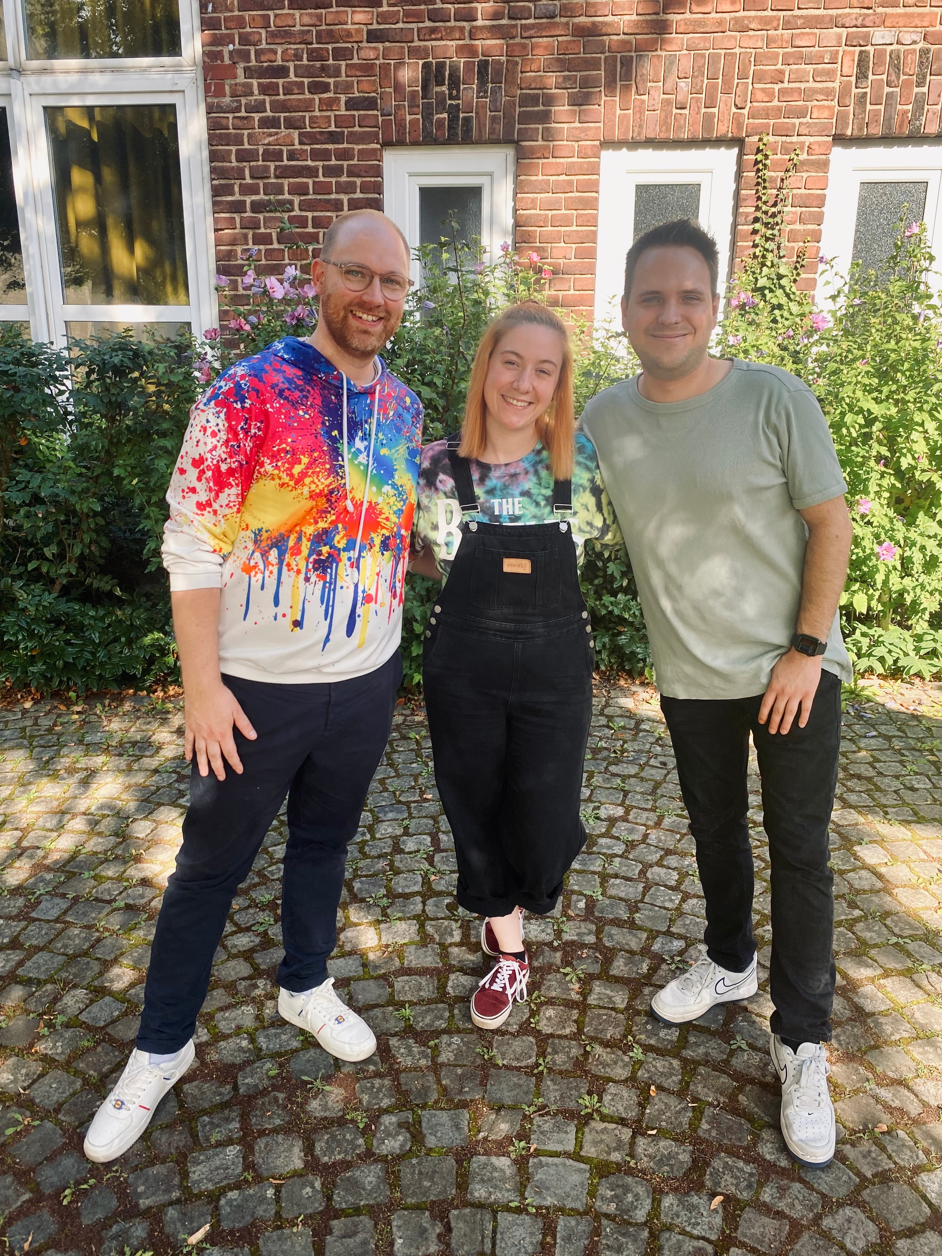 Drei Personen stehen lächelnd vor einer Hauswand