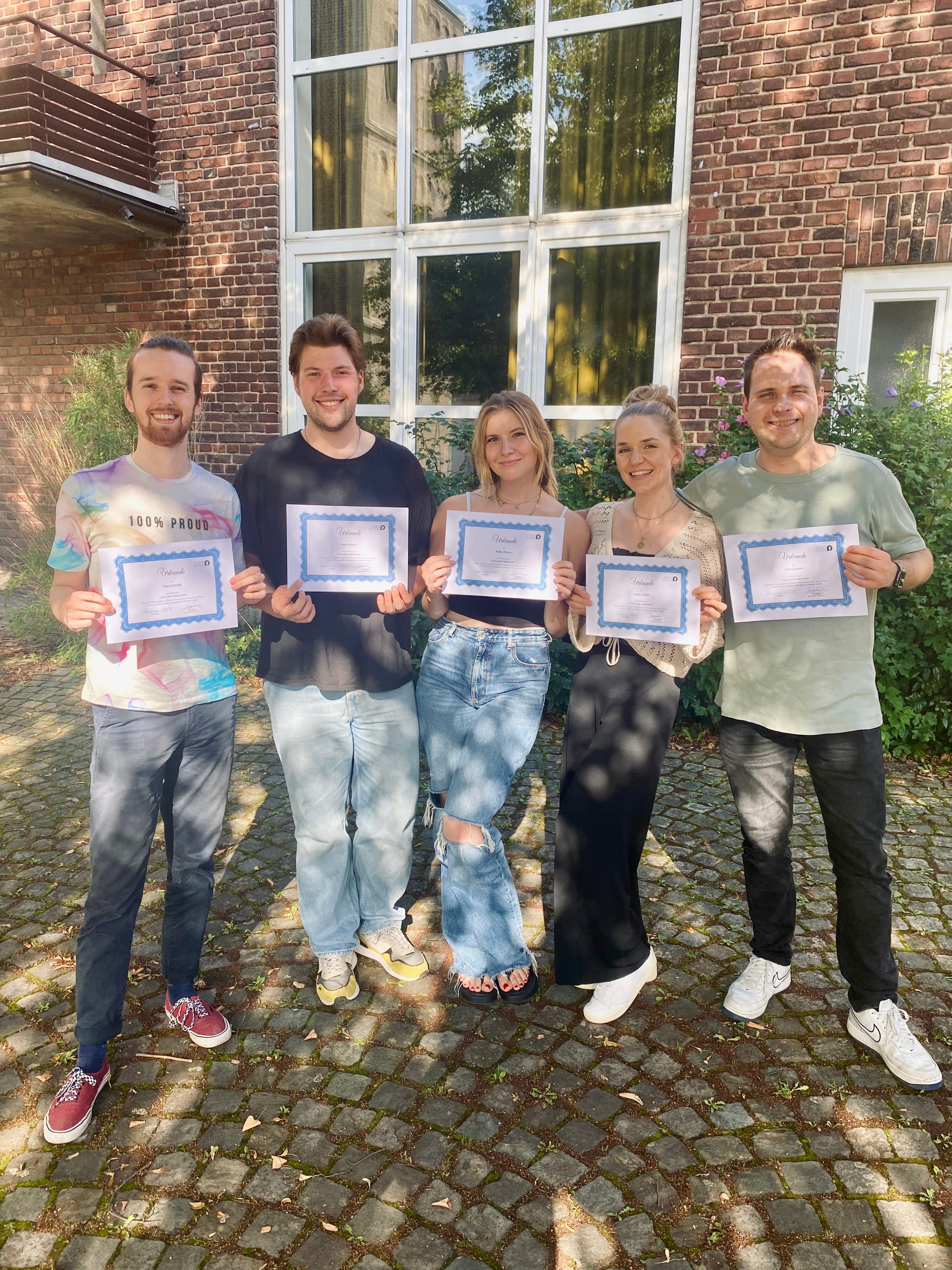 5 Personen stehen mit einer Urkunde in der Hand nebeneinander lächelnd vor einer Hauswand